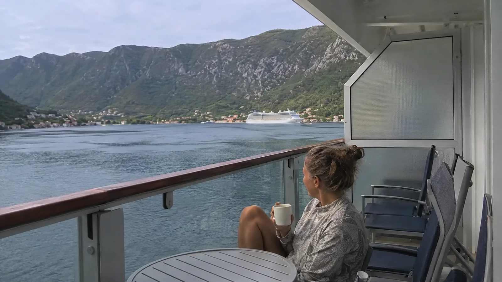 Woman sitting on a cruise ship balcony overlooking another ship and a mountainous harbor