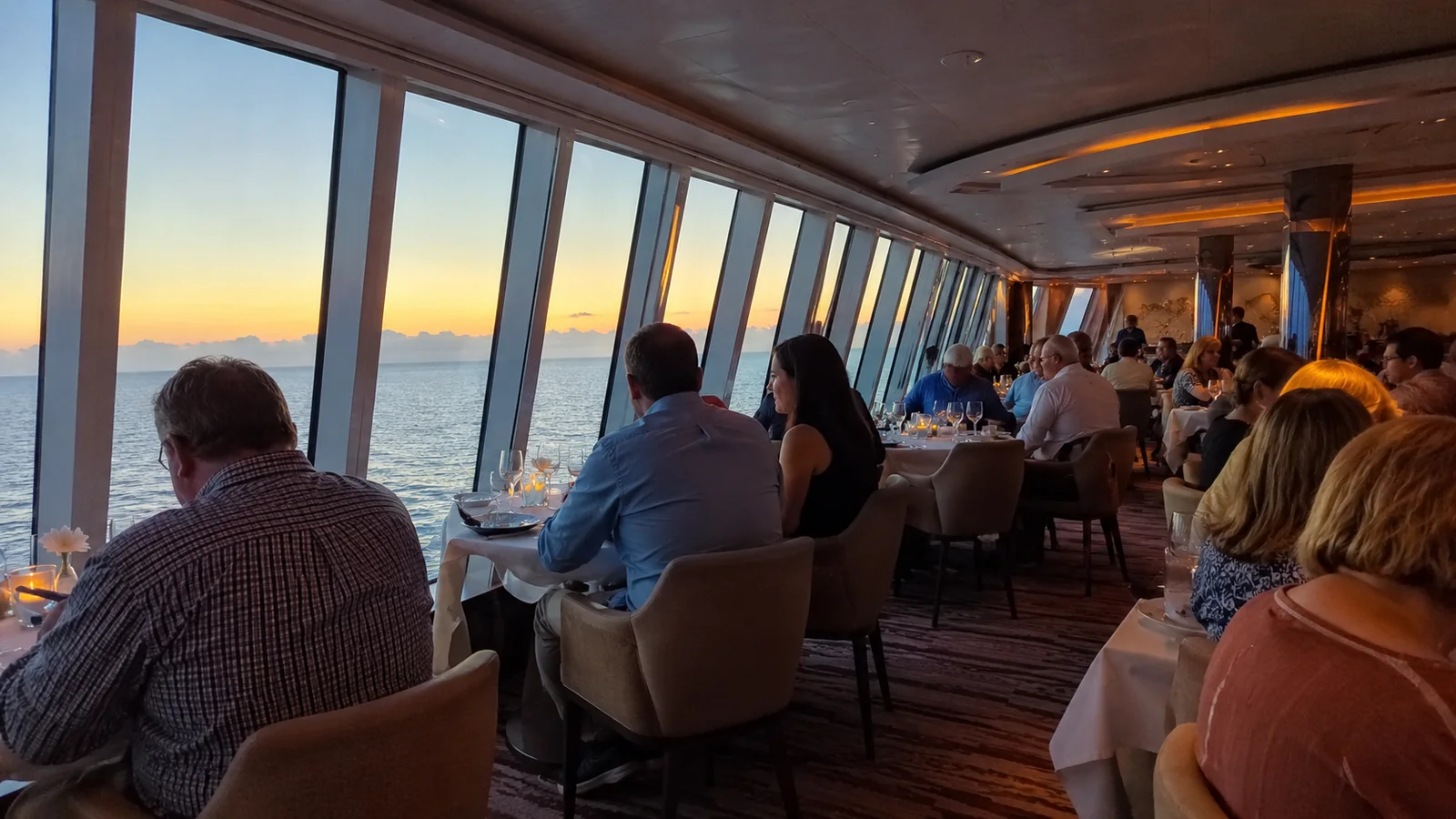 Passengers seated beside floor-to-ceiling windows in a cruise ship dining area at sunset