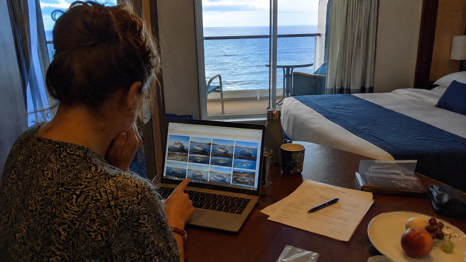 Woman comparing cruise ship photos on a laptop at a cabin desk beside an ocean-view balcony