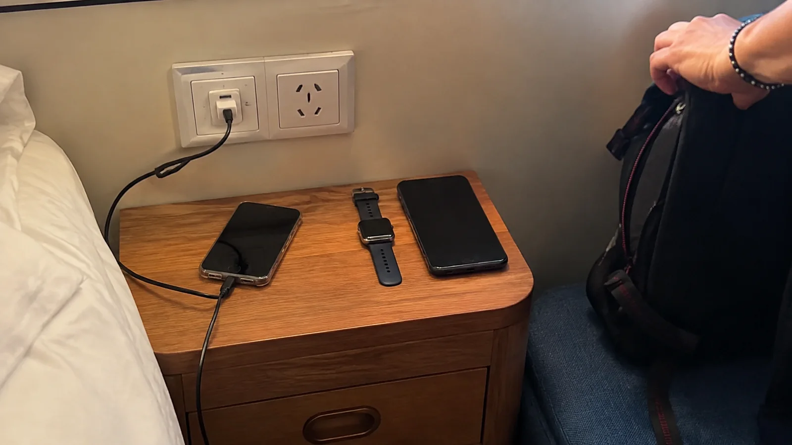 Phone charging beside a watch and tablet on a cabin bedside table.