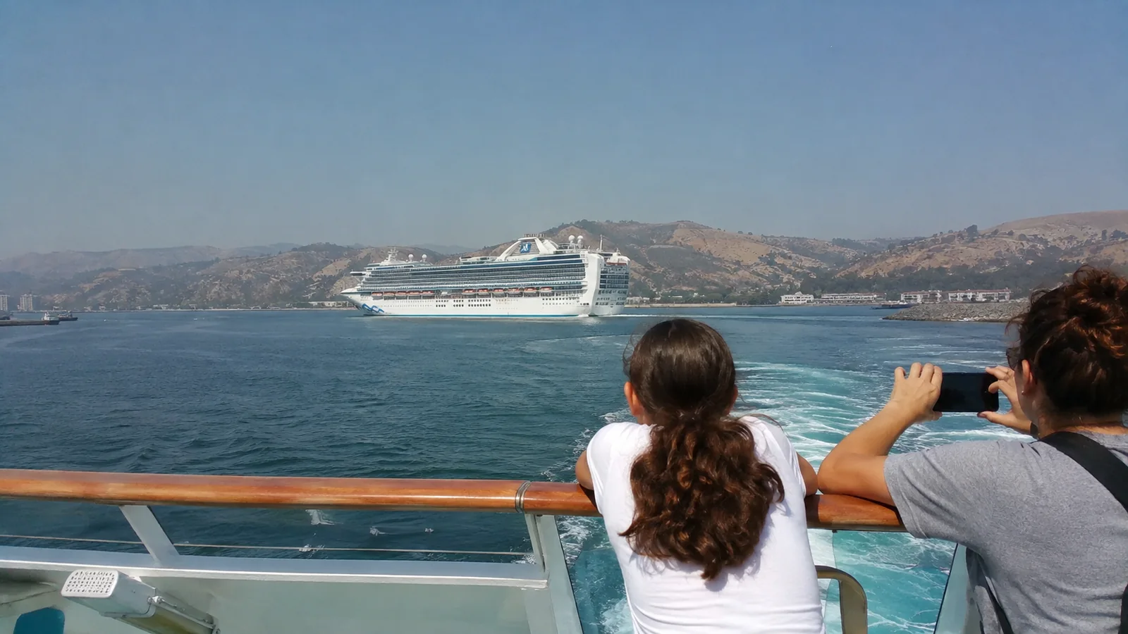 Passengers watching a cruise ship near a hilly coastline from an open deck.