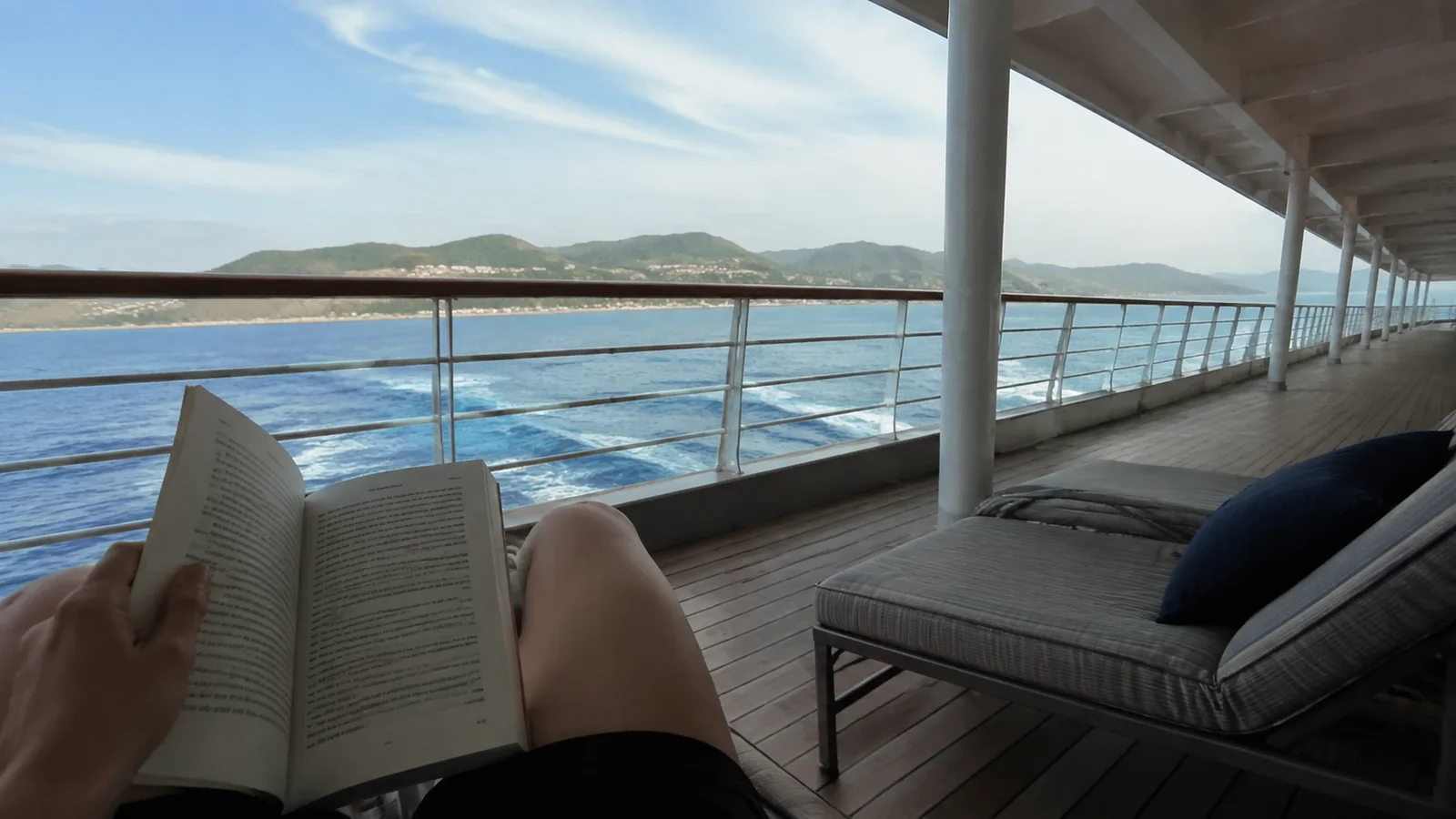 Passenger reading a book on a shaded deck overlooking the sea and hills