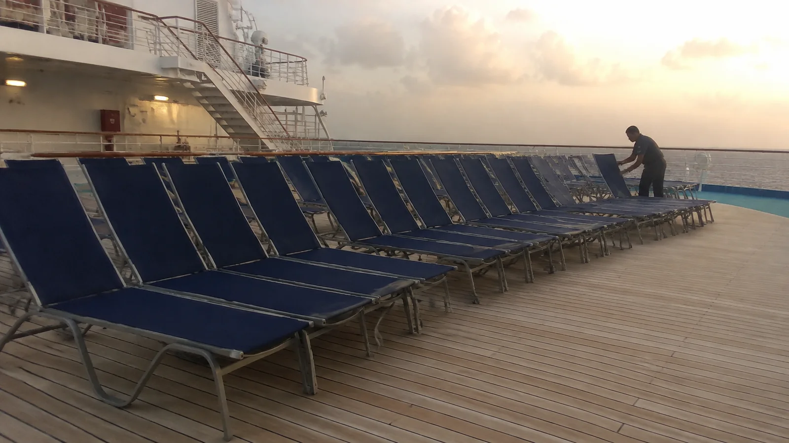 Crew member arranging rows of blue deck loungers at sunrise.