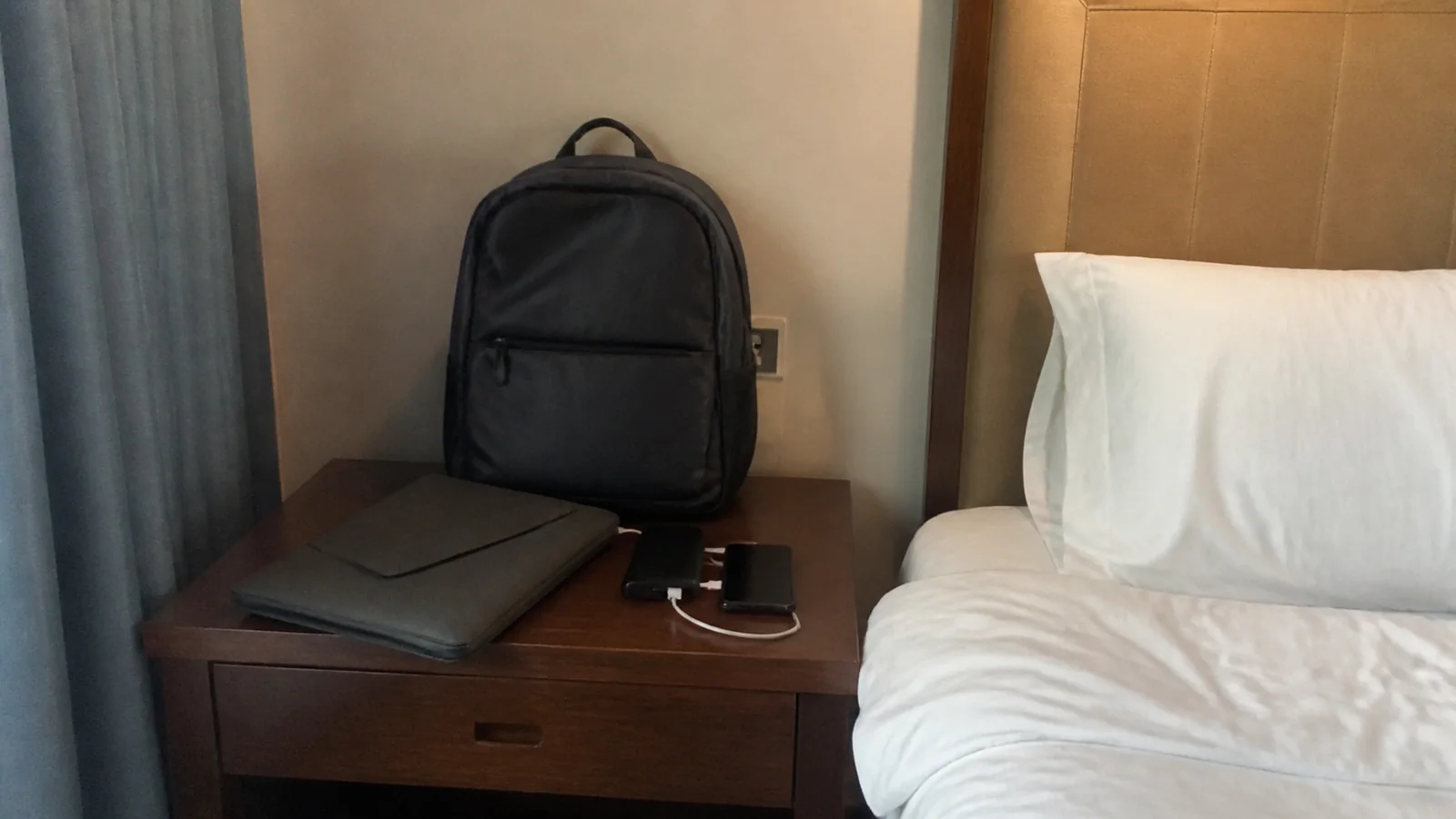 Backpack, closed laptop, and phone on a bedside desk.