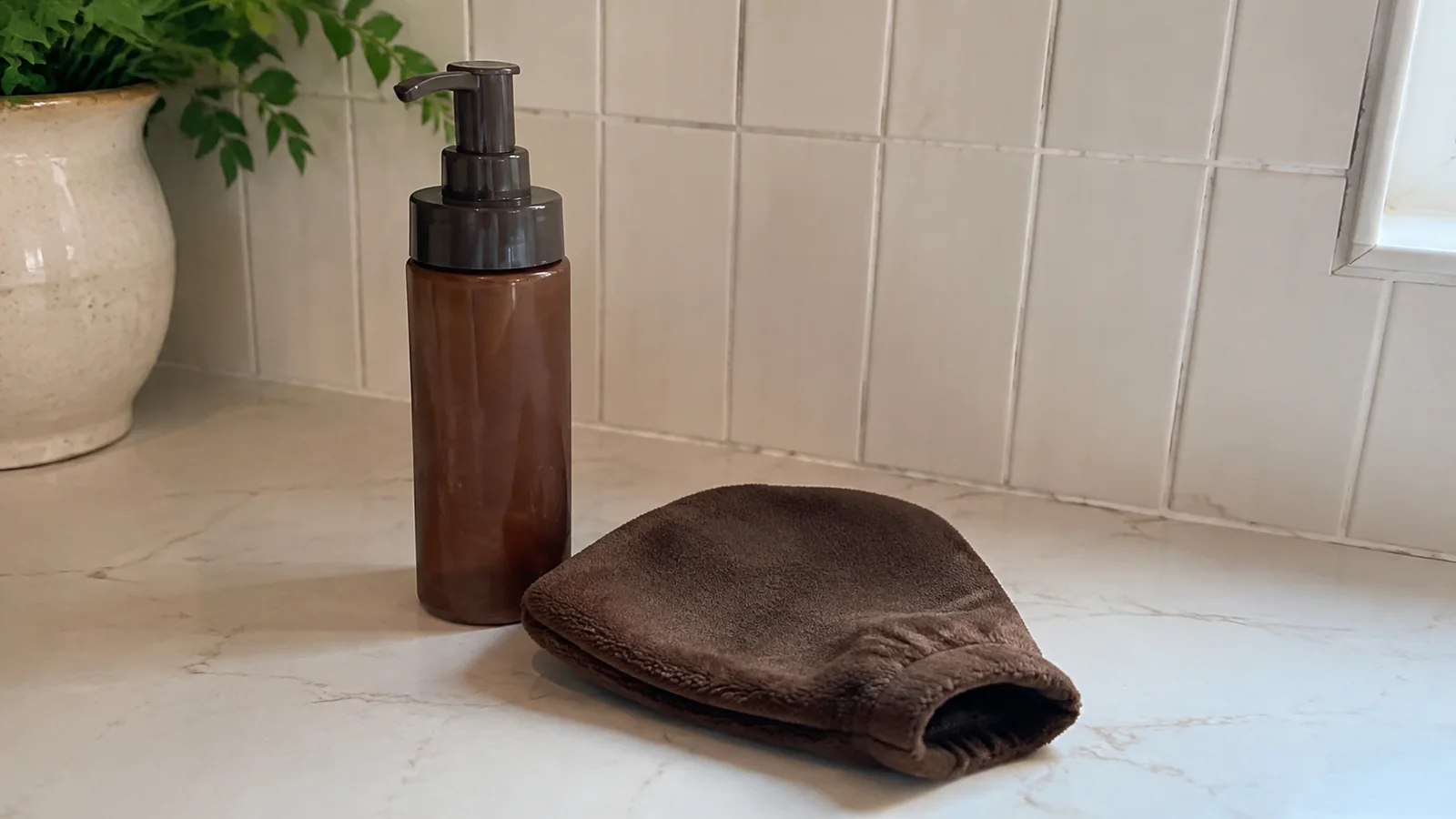 Amber pump bottle and brown applicator mitt on a bathroom counter.