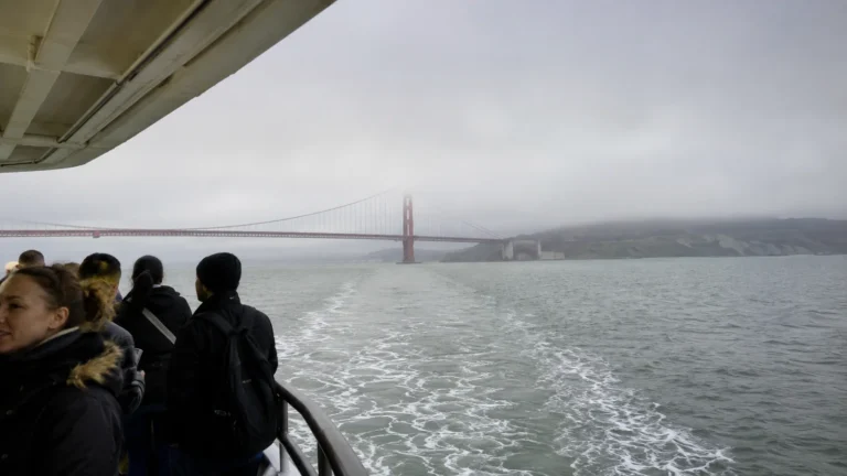 Passengers watching the Golden Gate Bridge recede into fog from a ship's deck.