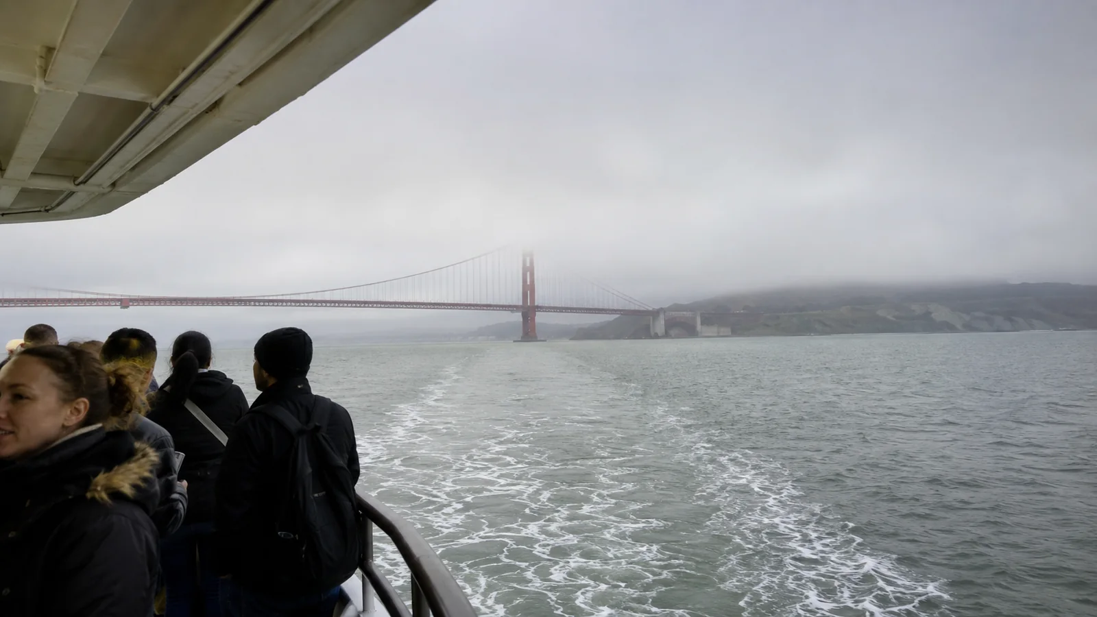 Passengers watching the Golden Gate Bridge recede into fog from a ship's deck.