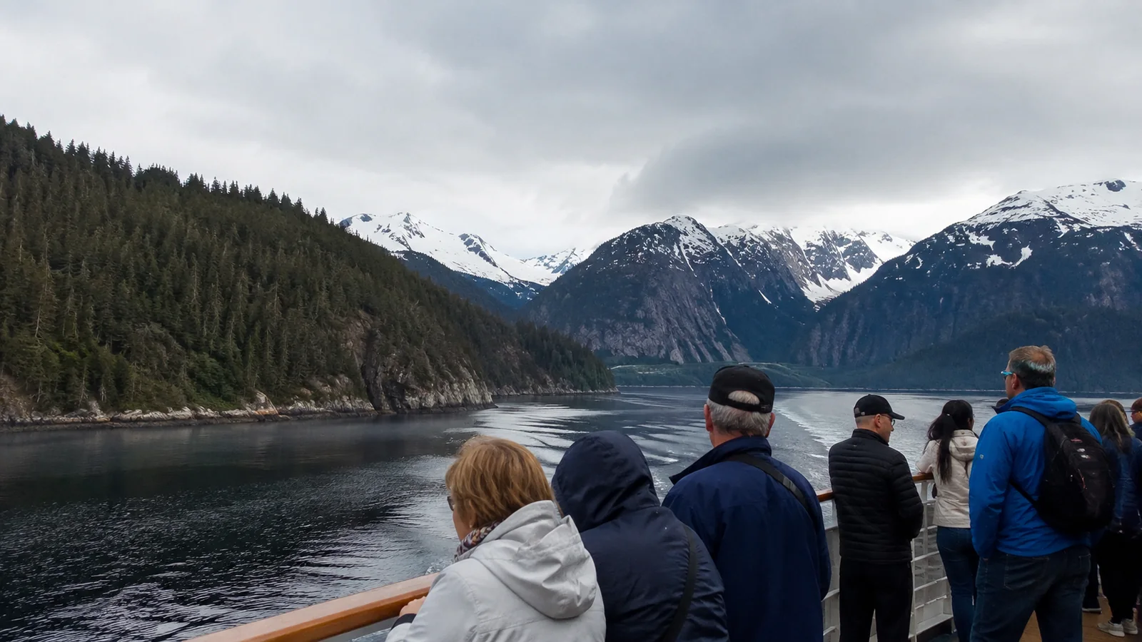 Passengers viewing forested shores and snowy mountains from a cruise ship.