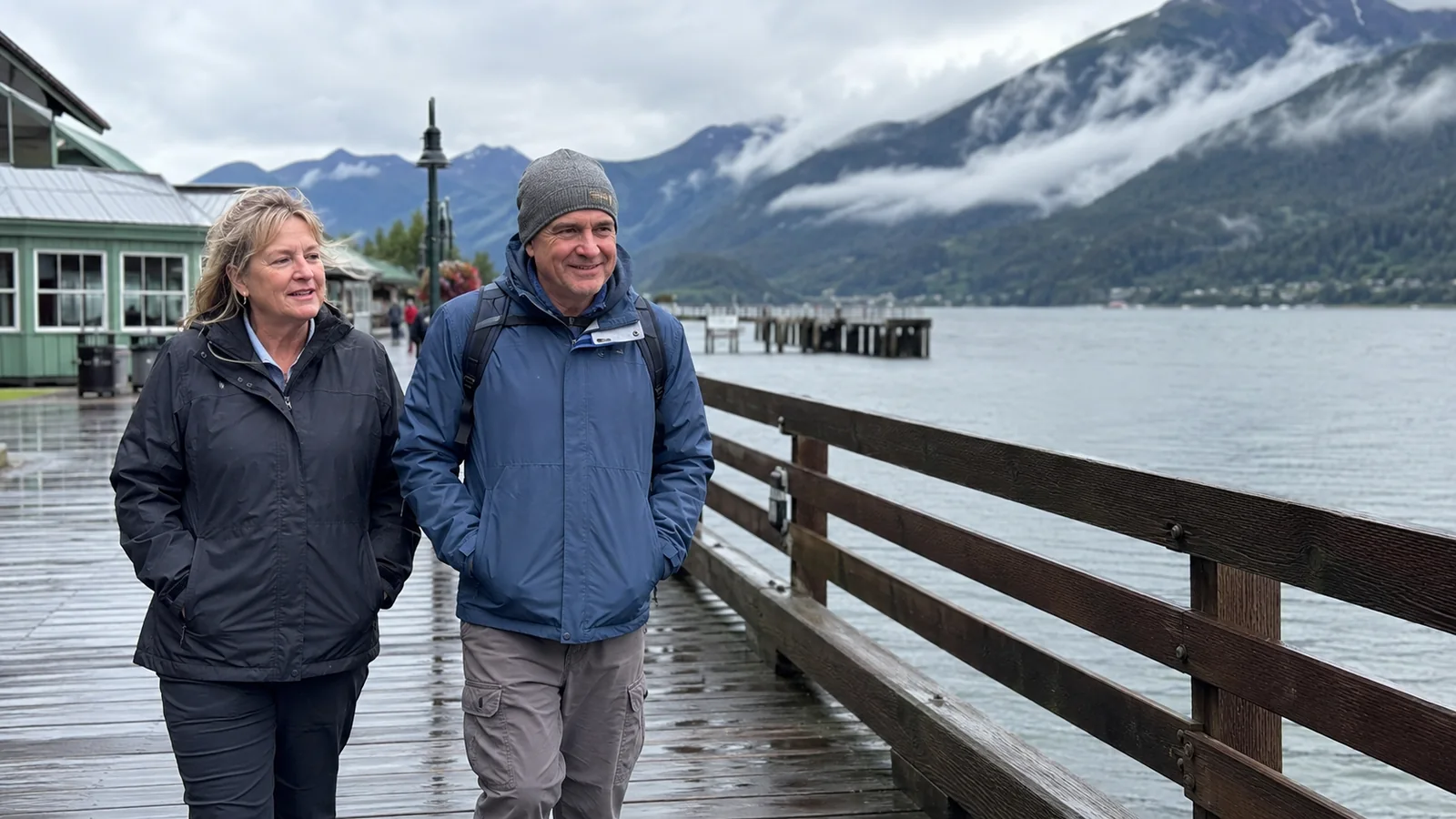 Two travelers wearing jackets walk beside a misty mountain waterfront.