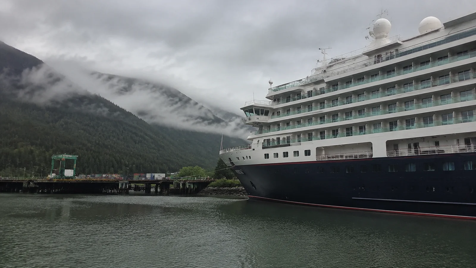 Cruise ship docked below mist-covered forested mountains.