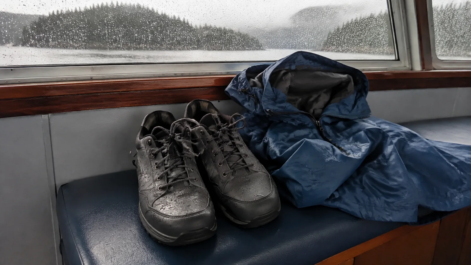Hiking shoes and a blue rain jacket drying beside a rain-speckled ship window.