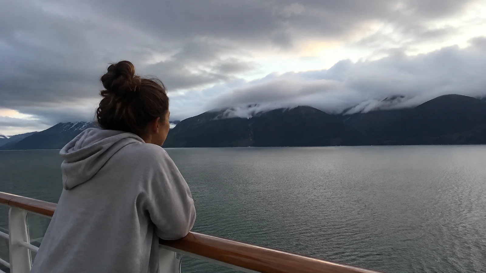 Woman leaning on a ship railing above calm water and cloud-covered mountains.