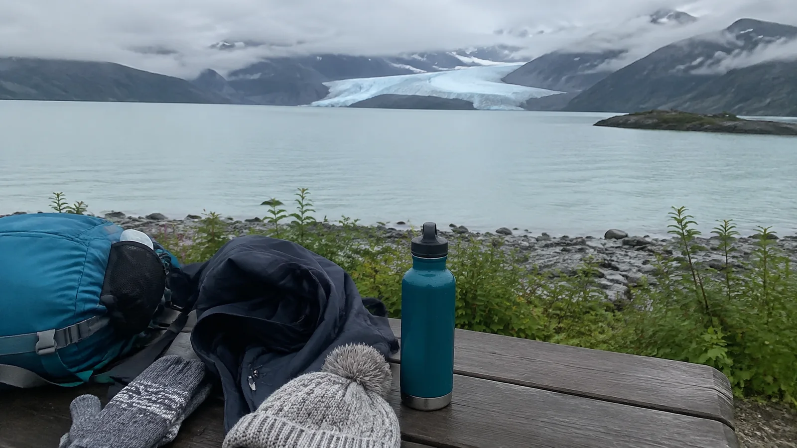 Backpack, knit hat, gloves, and water bottle on a bench overlooking a glacial bay