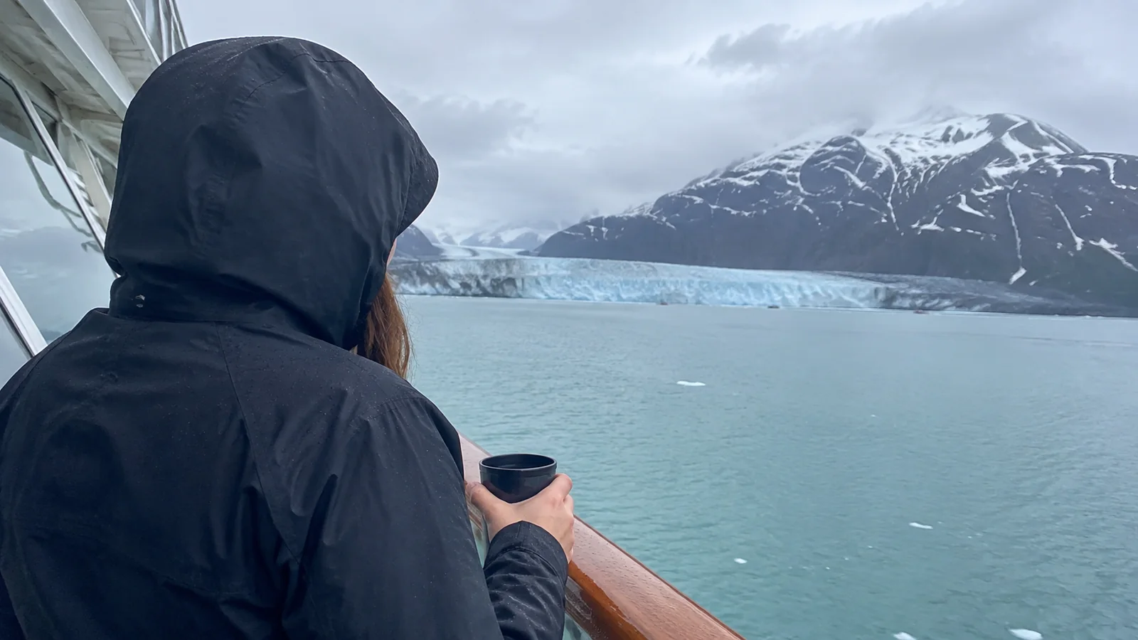 Hooded passenger holding a mug at a cruise ship railing with a glacier ahead