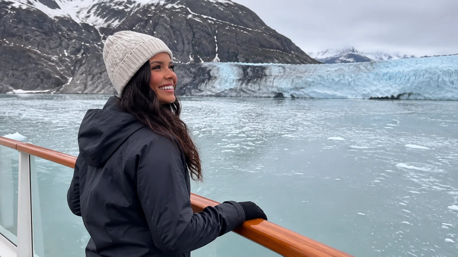 Woman in a knit hat standing at a cruise ship railing with a glacier behind her