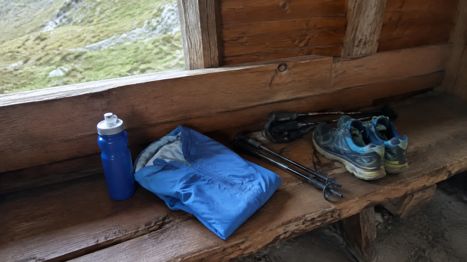 Hiking shoes, poles, rain jacket, and water bottle rest on a wooden bench.