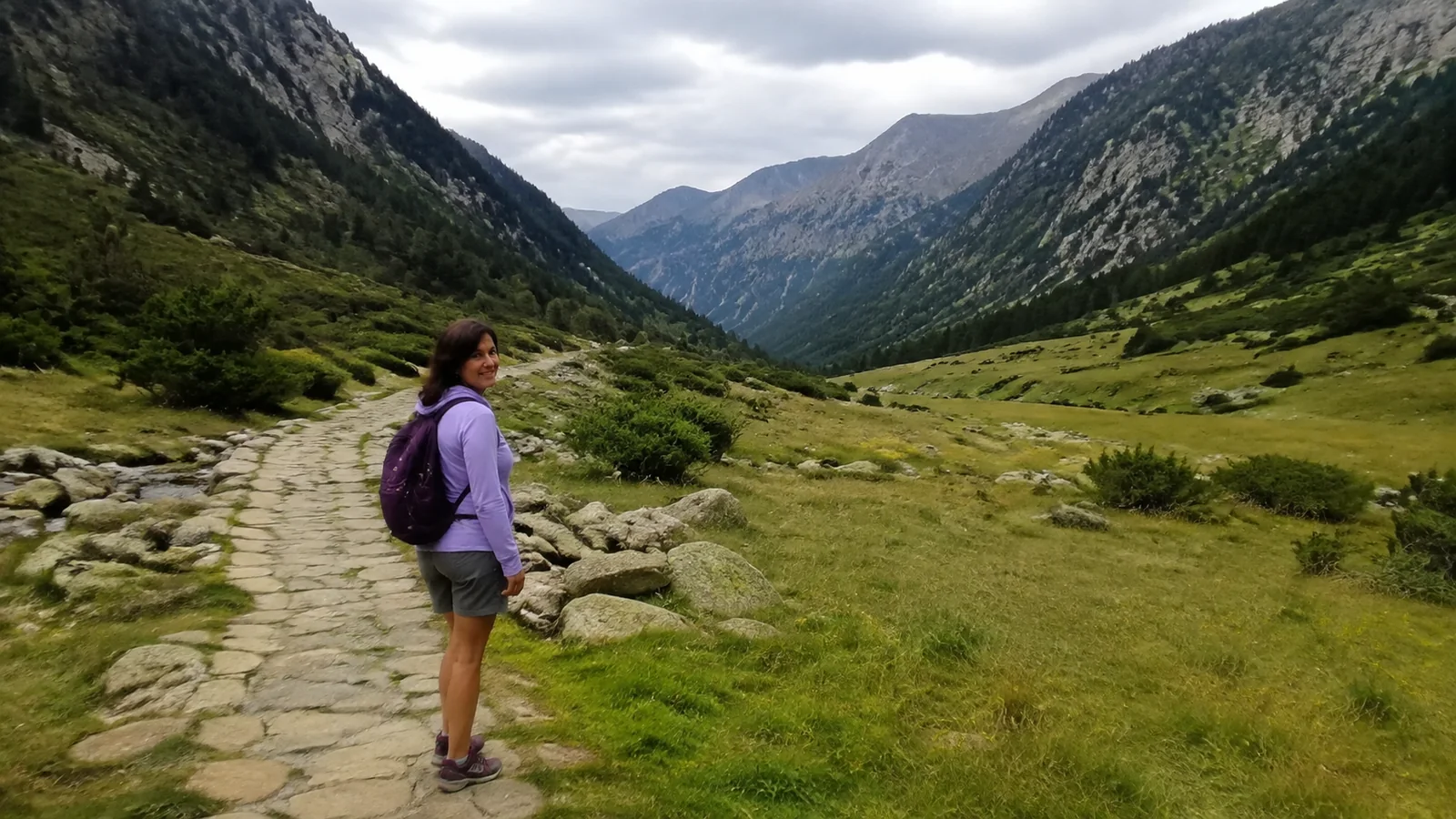 Hiker stands on a stone path through a broad green mountain valley.
