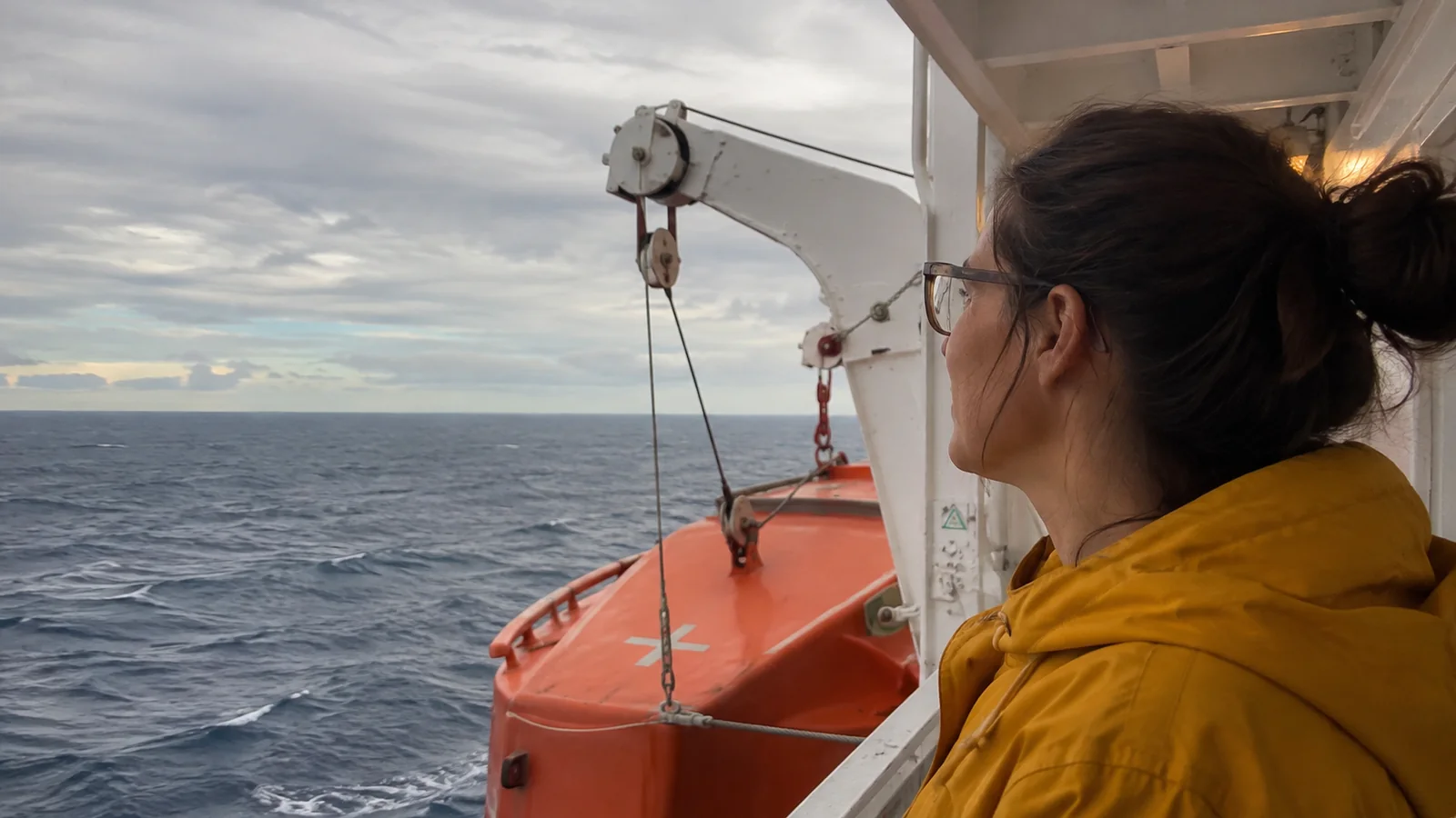 Passenger looking past an orange lifeboat toward the ocean.