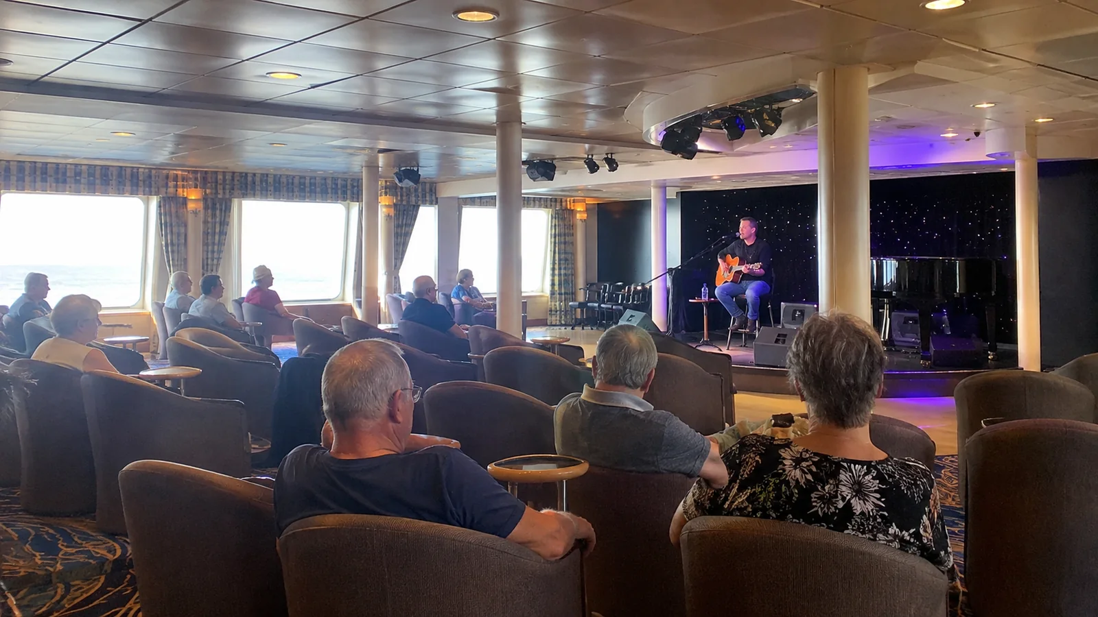 Passengers seated in a ship lounge watching a musician perform.