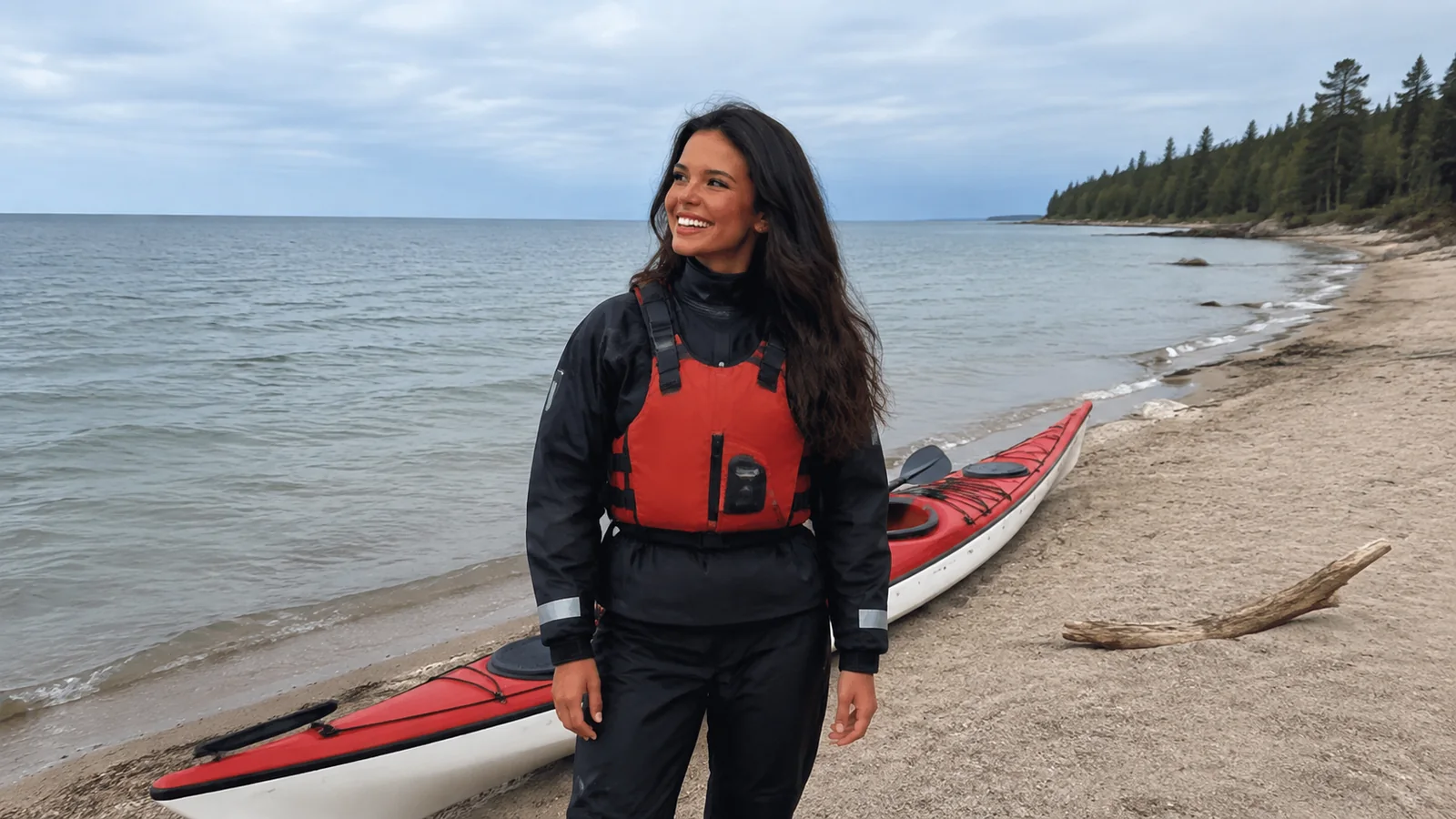 Woman wearing a red life jacket stands beside a kayak on a beach.