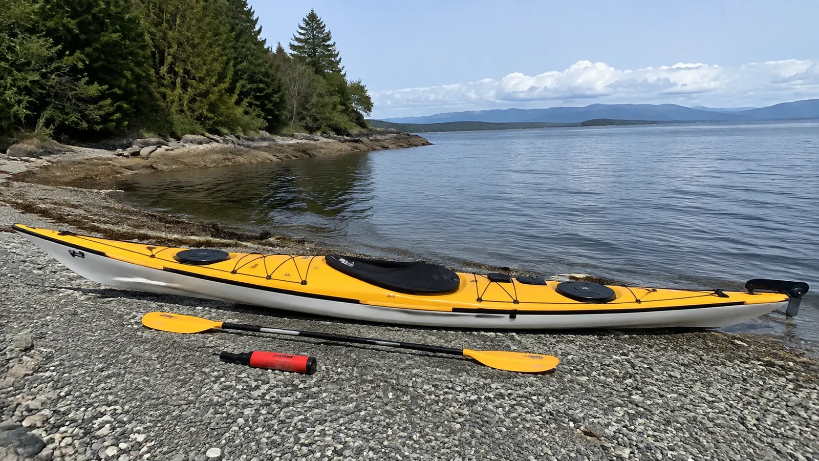 Yellow sea kayak, paddle, and small pump rest on a pebbly shore.