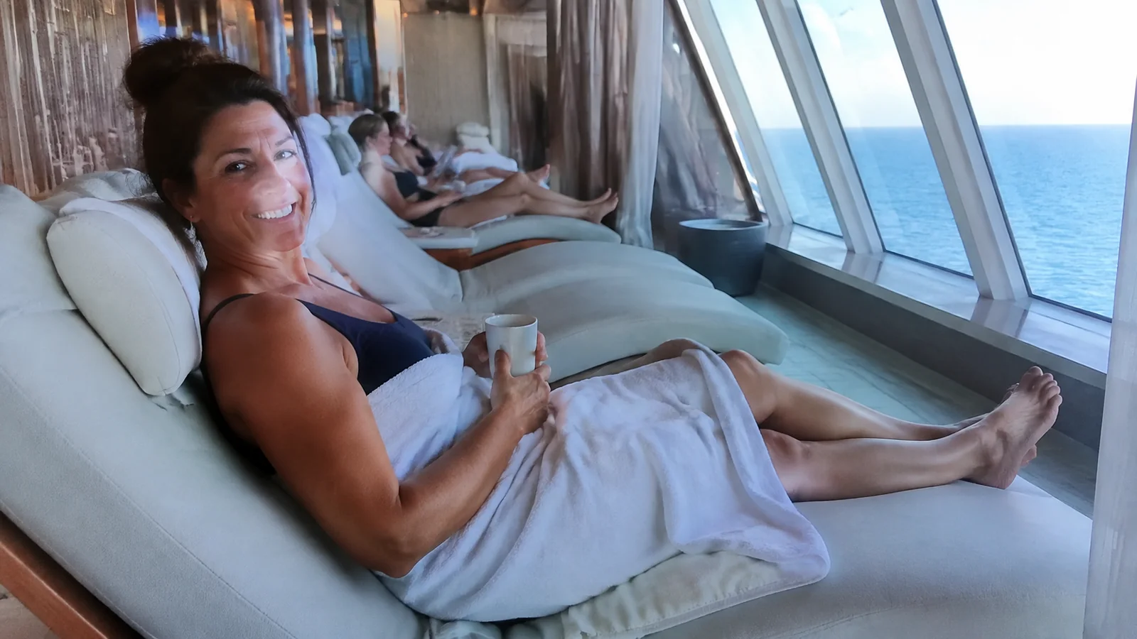 Passengers recline on loungers beside large windows overlooking the ocean.
