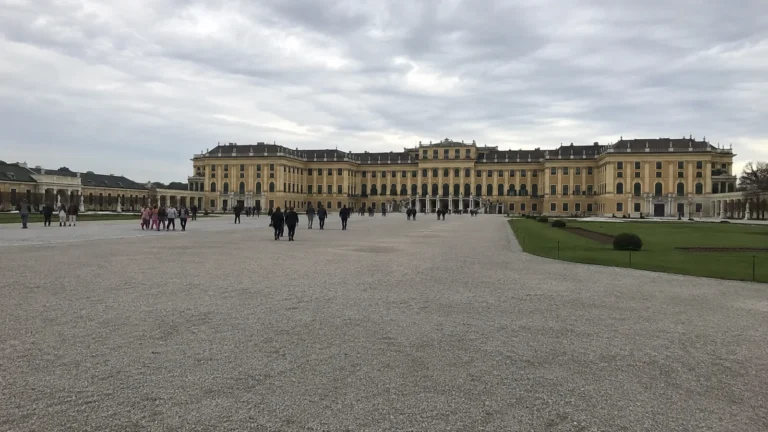 Visitors cross a broad courtyard in front of a yellow palace.