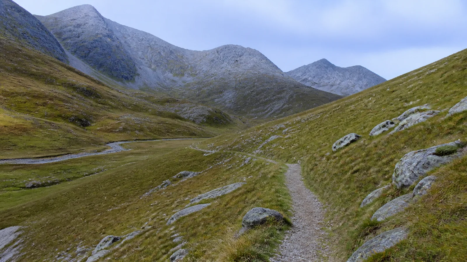 Narrow hiking trail crosses a green mountain valley beside a stream.