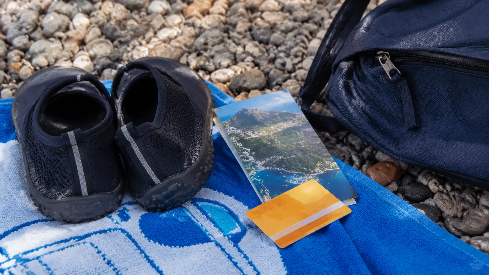 Black water shoes on a blue towel beside a folded map and backpack on a pebble beach