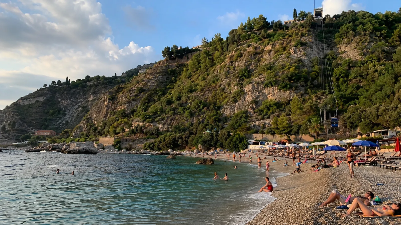 Crowded pebble beach beneath steep green cliffs beside clear blue water