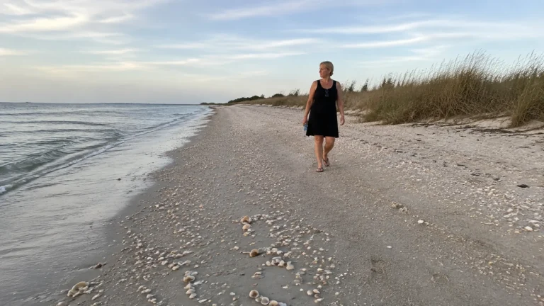 Woman walking barefoot along a shell-covered beach beside grassy dunes.