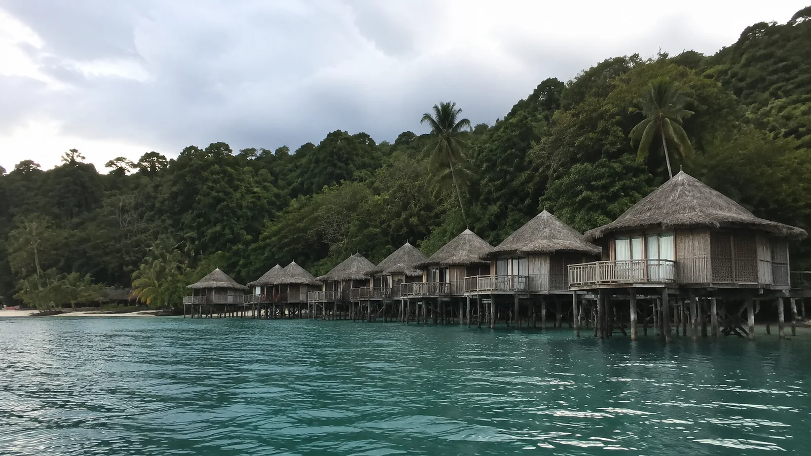 Row of rustic overwater bungalows backed by a densely wooded hillside.