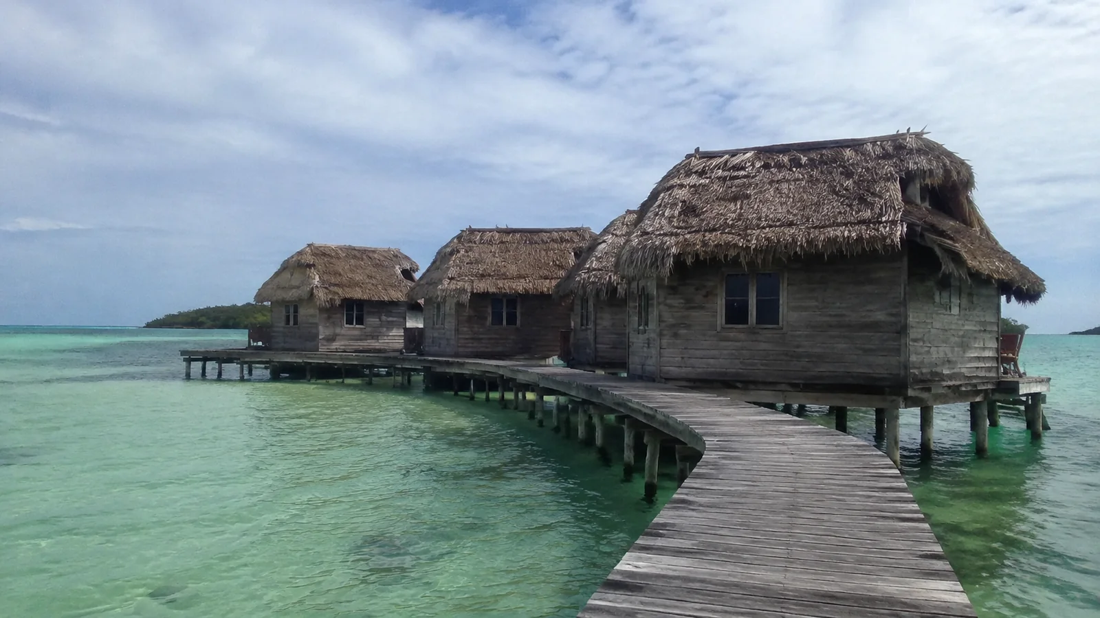 Curving boardwalk linking rustic thatched bungalows over pale turquoise water.