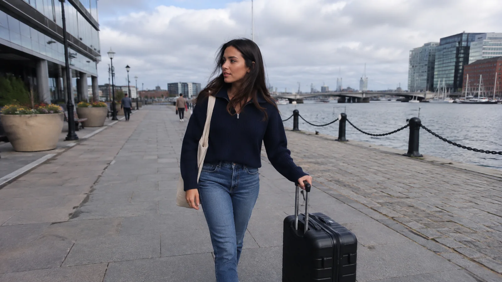 Woman wheeling a suitcase along an urban waterfront promenade.