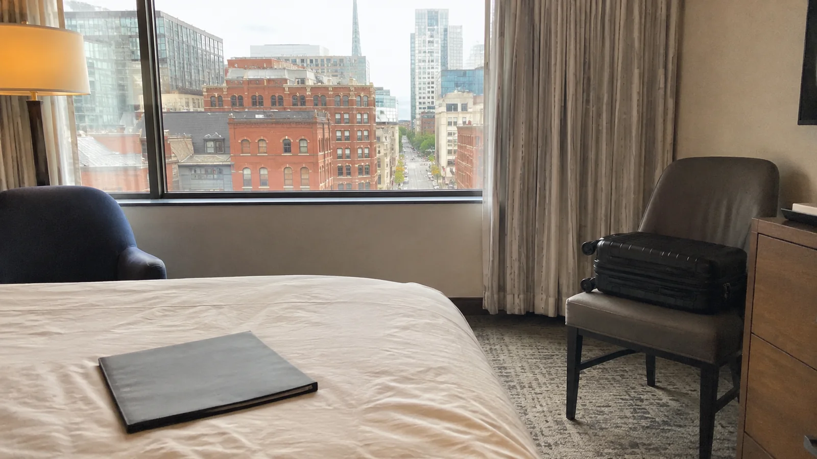 Hotel room with a suitcase on a chair and city buildings outside the window.