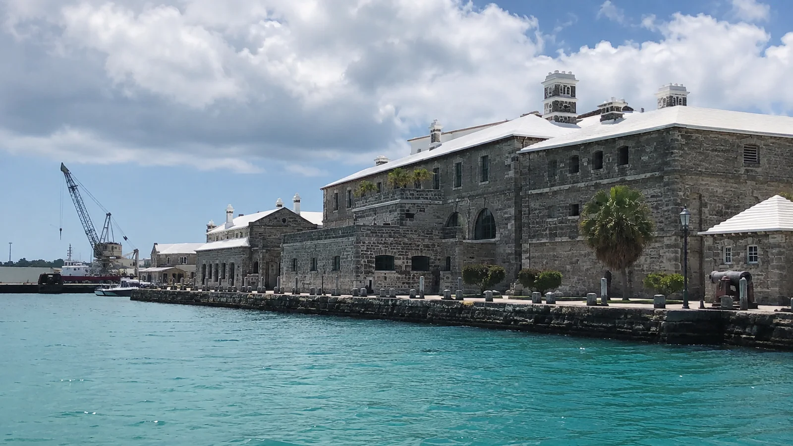 Historic stone dockyard buildings beside clear turquoise harbor water.