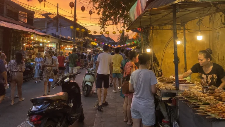 Crowded outdoor market street at dusk with food stalls and scooters