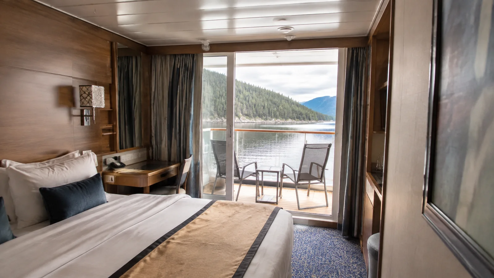 Ship cabin with a bed and balcony facing forested mountains.