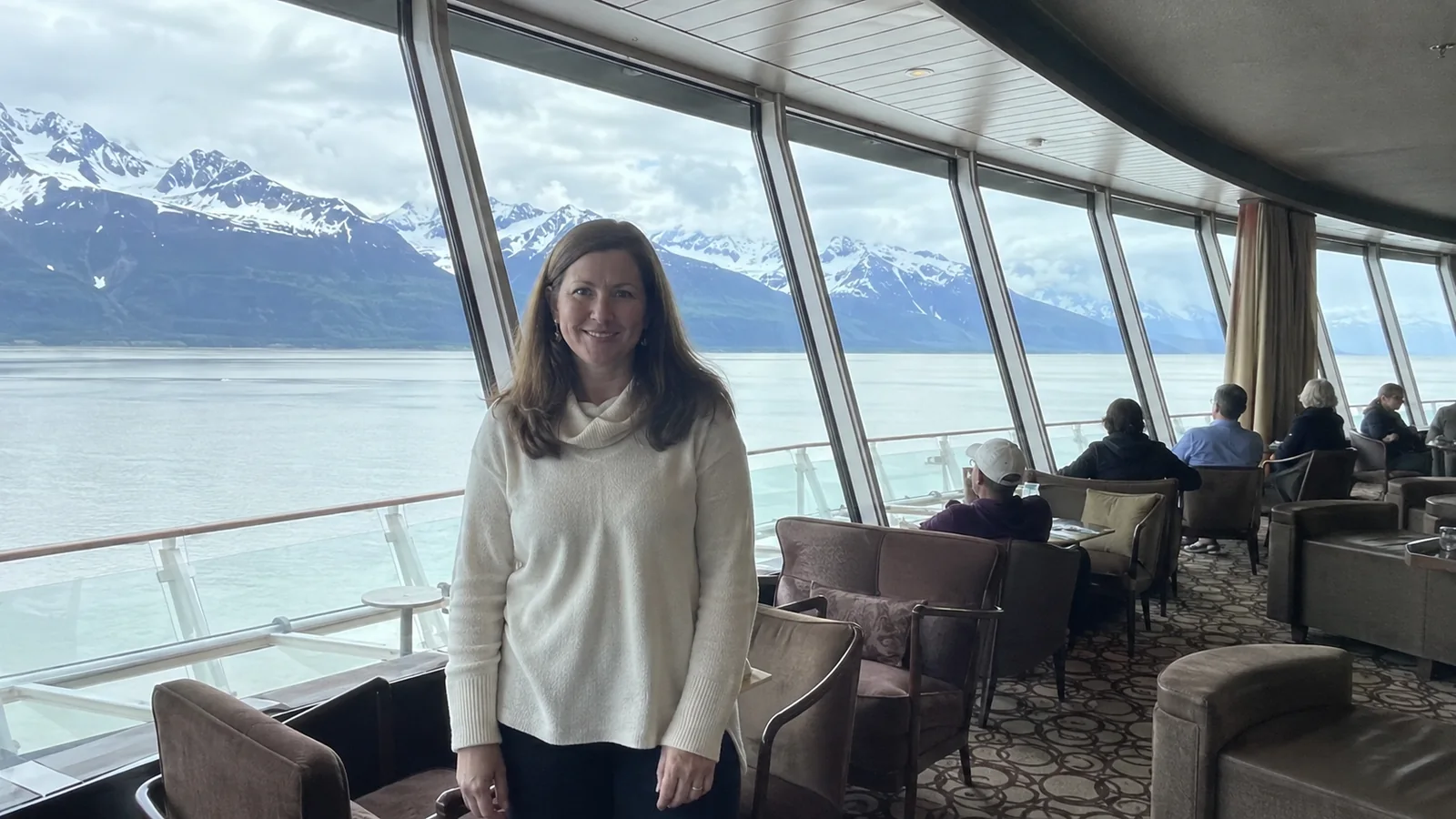 Woman standing in a panoramic ship lounge with snowy mountains outside.