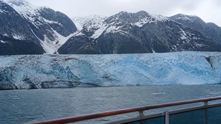 Blue glacier meeting the sea below steep snow-covered mountains.