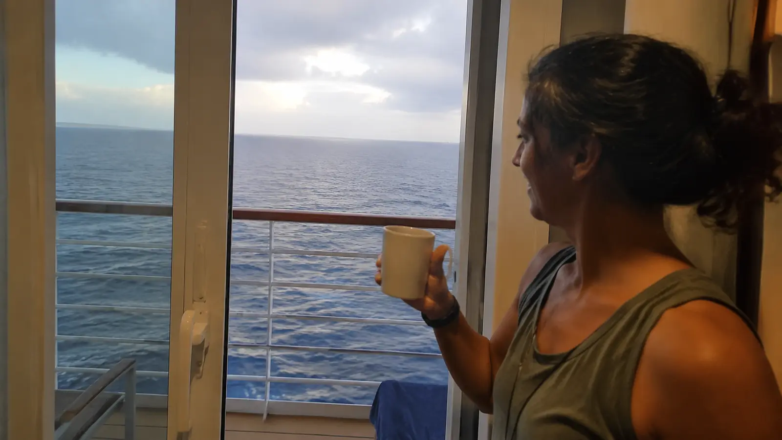 Woman holding a mug beside cruise cabin balcony doors overlooking the ocean