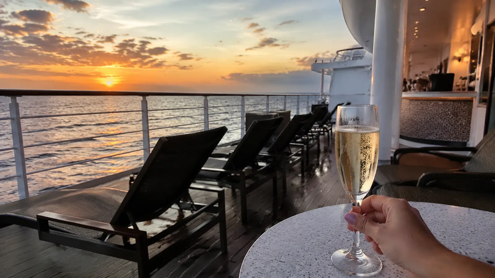 Hand holding a wine glass beside empty cruise ship deck chairs at sunset