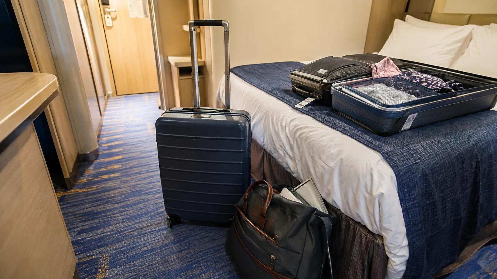 Suitcases and a tote bag arranged beside a bed in a cruise cabin.