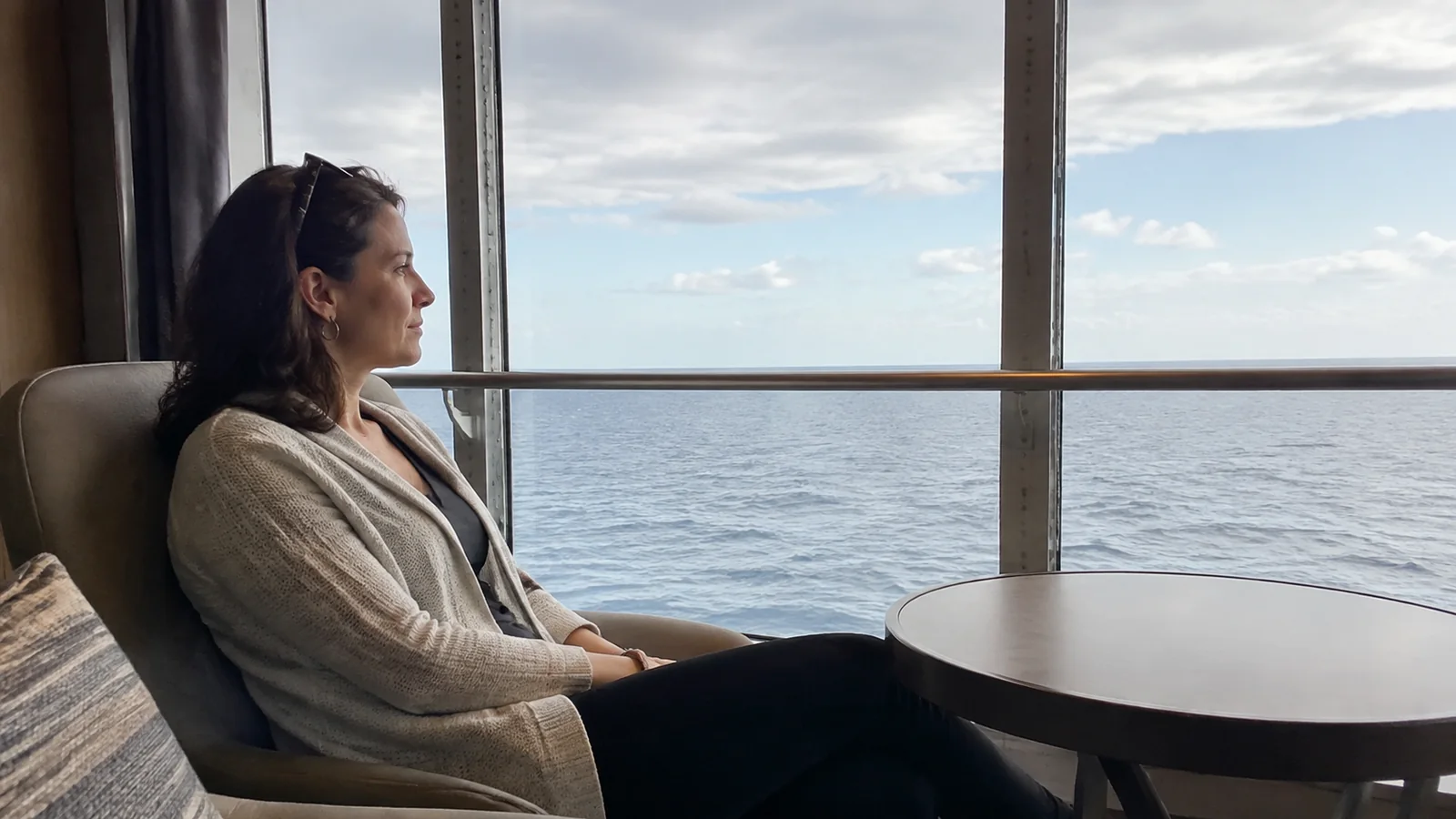 Woman sits beside a large window facing the level sea horizon.