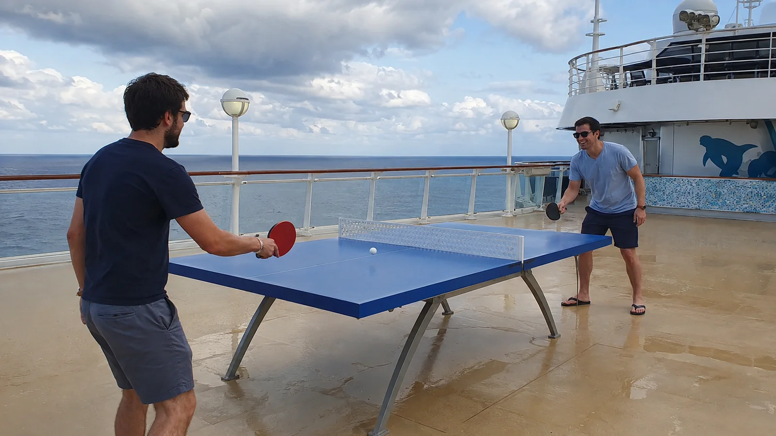 Two passengers playing table tennis on an open ship deck.