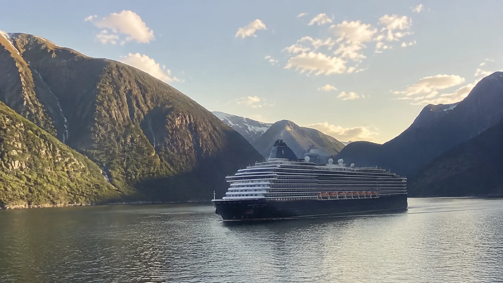 Cruise ship sailing between steep mountains in a sunlit fjord.