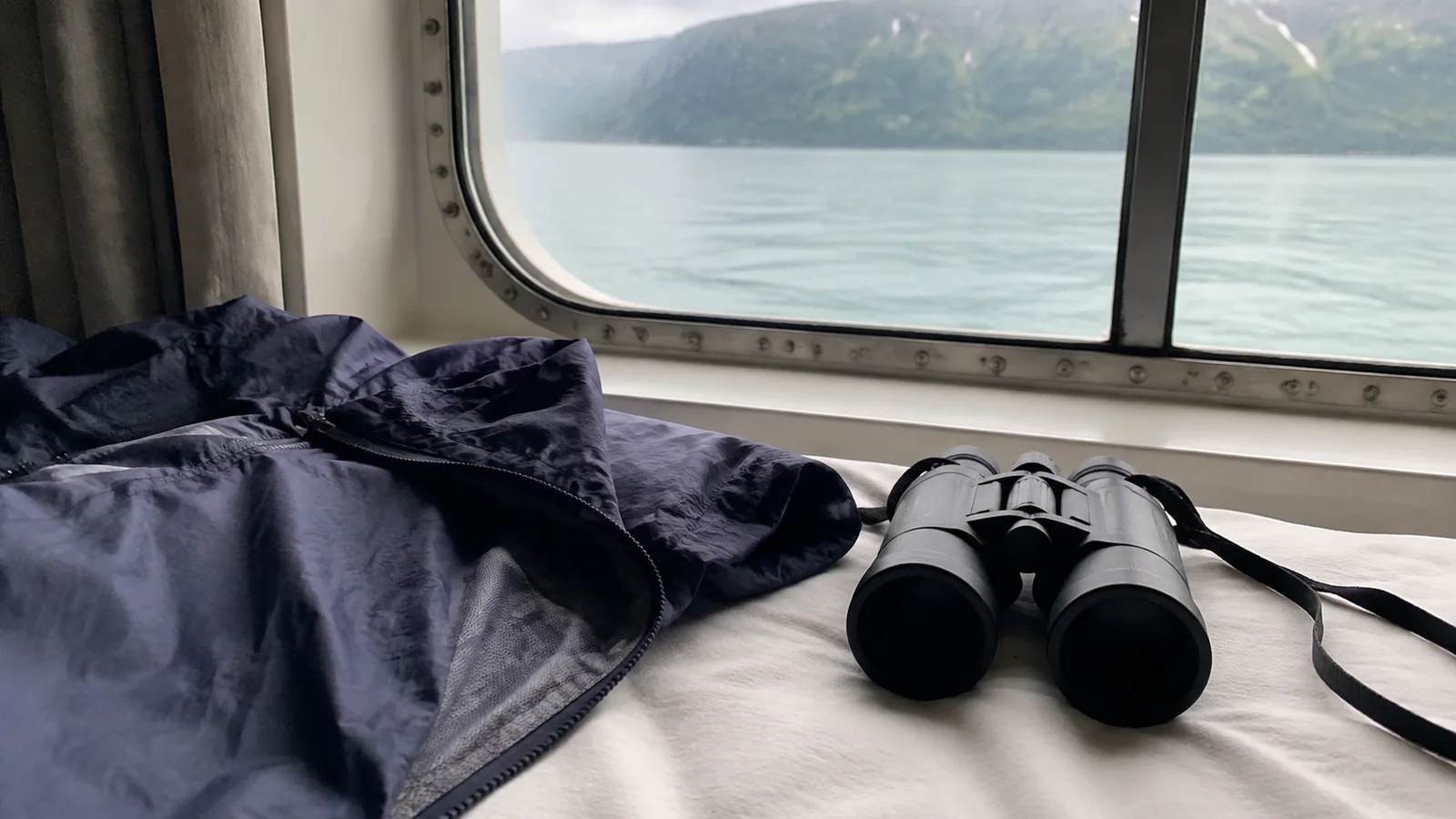 Binoculars and a rain jacket beside a ship window overlooking a fjord.