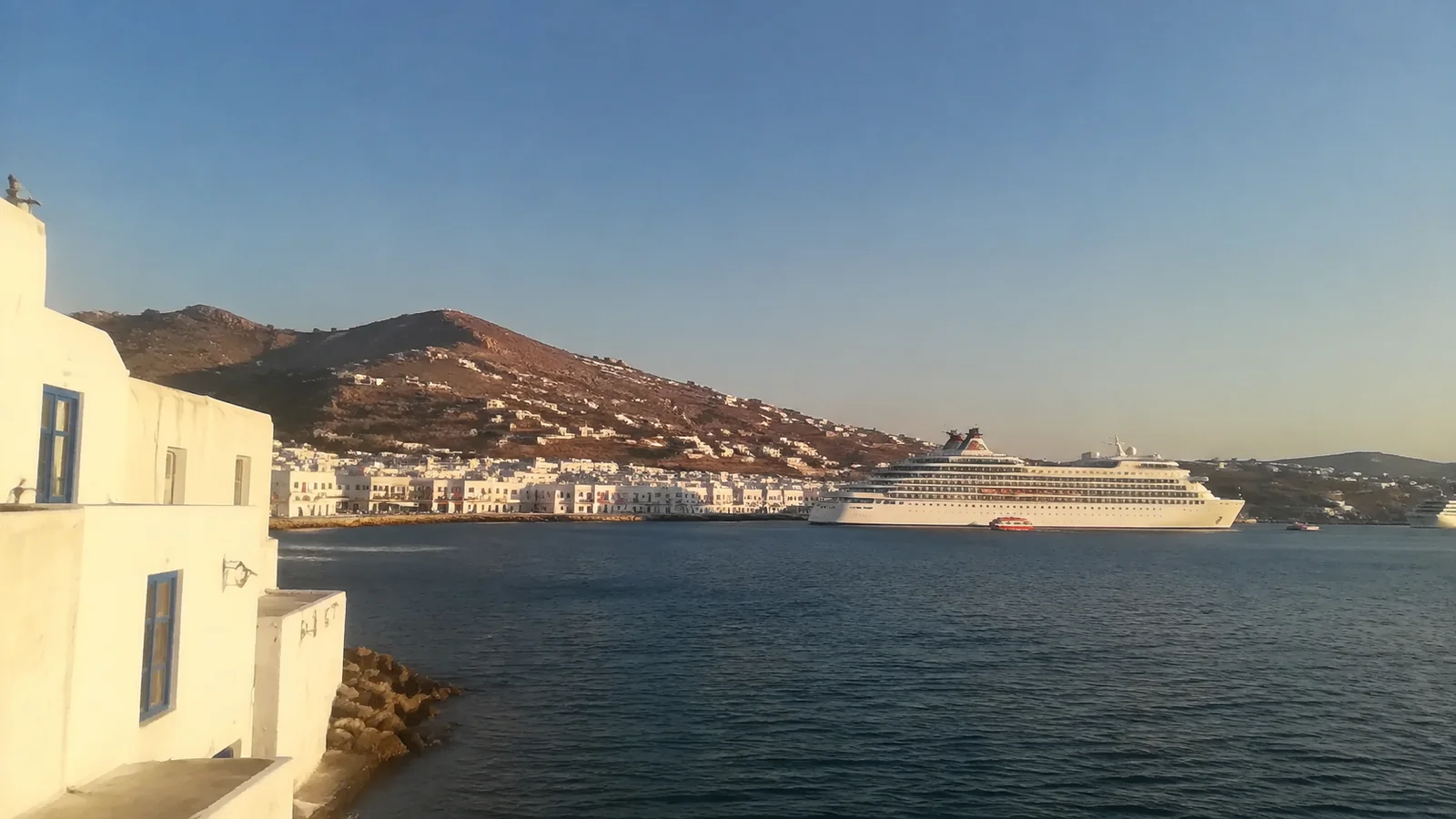 Cruise ship passing a hillside harbor town in warm evening light.