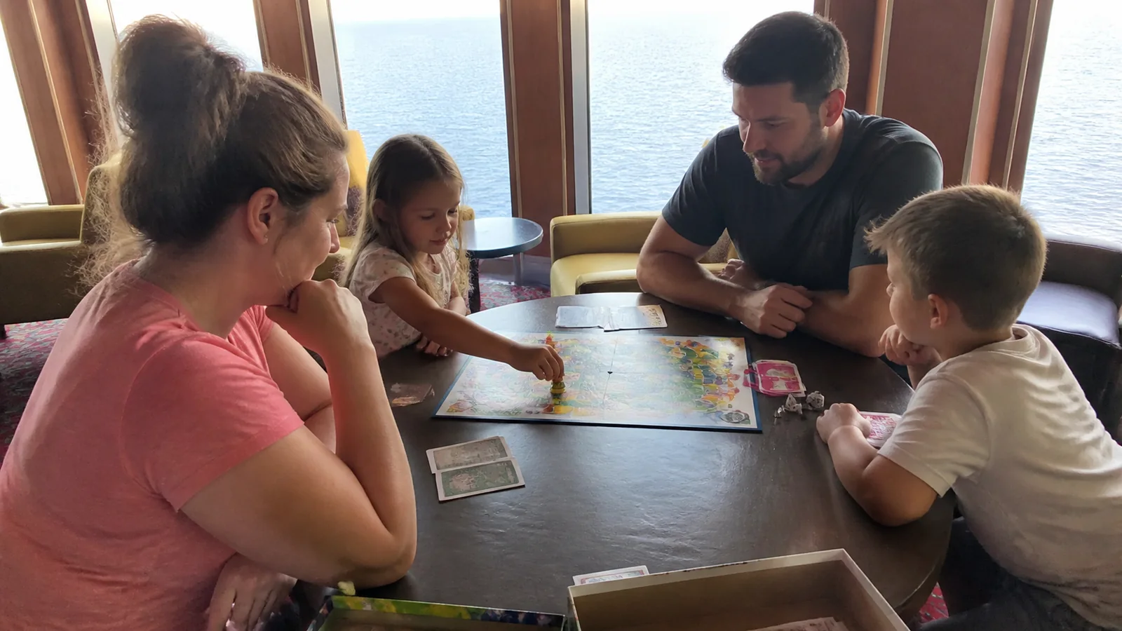 Family playing a board game beside a ship's ocean-view windows.