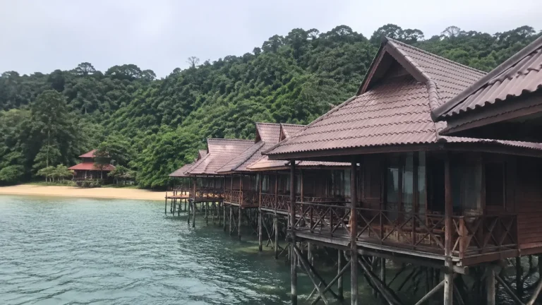 Timber chalets on stilts beside calm water and a forested tropical hillside.